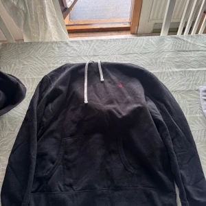 Svart hoodie från Polo Ralph Lauren - Svart hoodie från Polo Ralph Lauren med liten röd logga broderad på bröstet. Tröjan har huva med vita snören och en stor magficka framtill. Perfekt för en avslappnad stil.