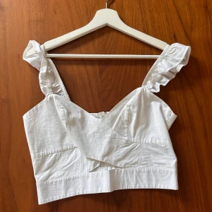 Vit croppad linnetopp med volangaxelband - Säljer en söt vit croppad linnetopp med volangprydda axelband och omlott-detalj framtill. Toppen har dragkedja i ryggen och är perfekt för varma dagar.
