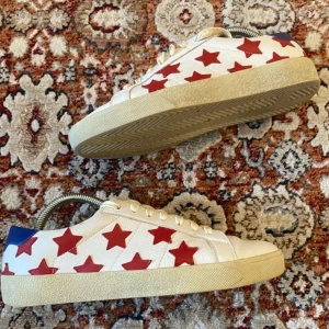 Vita sneakers med röda stjärnor från Saint Laurent - Säljer ett par vita sneakers från Saint Laurent med röda stjärnor och blå detalj på hälen. Skorna har snörning och platt sula, perfekt för dig som vill sticka ut med en unik design.
