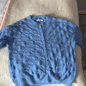 Blå kofta med mönster  - Säljer en ljusblå kofta från Jumperfabriken med fint stickat mönster och rund hals. Koftan har knappar framtill och ribbade muddar vid ärmslut och nederkant. Perfekt att slänga på sig över en topp!