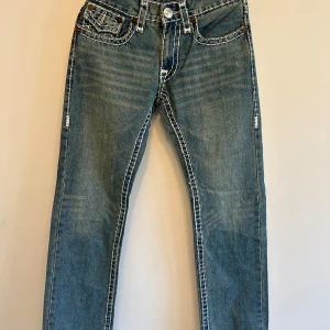 Blå jeans från True Religion med vita sömmar - Säljer ett par klassiska blå jeans från True Religion med tydliga vita kontrastsömmar och snygga bakfickor med lock och knapp. Jeansen har normal passform och raka ben. Perfekta för dig som gillar streetwear och vill sticka ut lite extra. 💕{Längd:103,5 cm, Innerbenslängd: 78,5cm}