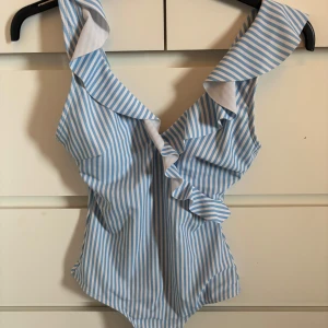 Blåvit randig baddräkt med volang - Supersöt baddräkt med blåvita ränder och volangdetaljer längs axelbanden och framtill. Perfekt för strand eller pool! Snygg och trendig modell med feminina detaljer.