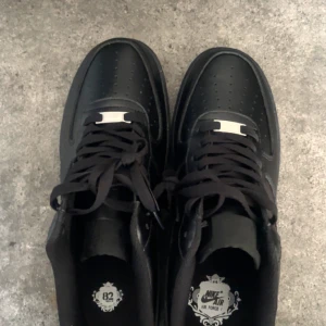 Svarta Nike Air Force 1 sneakers - Säljer ett par klassiska helsvarta Nike Air Force 1 sneakers med snörning och rund tå. Skorna har en platt sula och är tillverkade i syntetmaterial med diskret Nike-logga på sidan. Perfekta för dig som gillar stilrena och tidlösa sneakers. Helt nya.