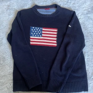 Mörkblå stickad tröja med amerikansk flagga från GANT - Säljer en mörkblå stickad tröja från GANT med en stor broderad amerikansk flagga på bröstet. Tröjan har rund halsringning och långa ärmar med ribbade muddar. Perfekt för dig som gillar klassisk och preppy stil.