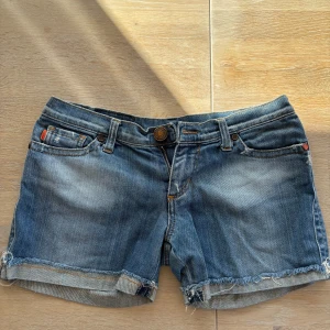 Blå jeansshorts från Frutta - Snygga blå jeansshorts från Frutta med råa kanter och klassisk femficksdesign. Shortsen har normal passform, låg midja och detaljerade sömmar. Perfekta för sommardagar och enkla att matcha med olika toppar.