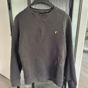 Mörkgrå sweatshirt från Lyle & Scott - Säljer en mörkgrå sweatshirt från Lyle & Scott med rund halsringning och klassisk gul logga på bröstet. Tröjan har långa ärmar och ribbade muddar vid ärmslut och nederkant. Perfekt för en avslappnad stil.