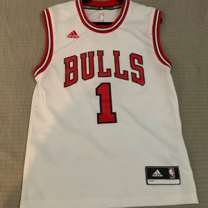 Chicago Bulls baskettröja Rose #1 - Säljer en vit Chicago Bulls baskettröja från Adidas med röd och svart kant, nummer 1 och namnet Rose på ryggen. Klassisk NBA-design med stora tryck och officiell logga. Perfekt för basketfans som vill sticka ut på match eller träning.