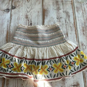 Vit kjol med broderade blommor - Söt vit kjol med smockad midja och volang. Kjolen har broderade gula och röda blommor med gröna blad längs nederkanten. Perfekt för en somrig look med boho-vibbar.