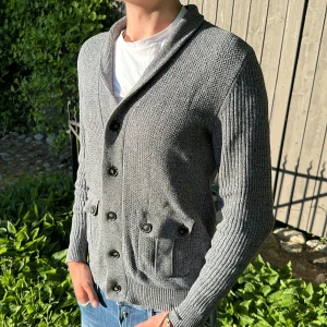 Grå stickad cardigan med knappar - Snygg grå stickad cardigan med flera svarta knappar framtill och två fickor med knappdetaljer. Cardiganen har lång ärm och ribbade partier på ärmar och baksida. Perfekt för lager-på-lager och passar till jeans.