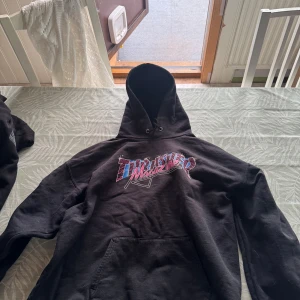 Svart hoodie från Thrasher Magazine - Säljer en svart hoodie från Thrasher Magazine med färgglad logga på bröstet. Klassisk modell med huva och känguruficka framtill. Perfekt för dig som gillar streetwear och skatekultur.