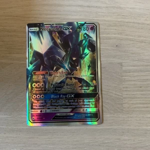 Necrozma GX Pokémonkort - Säljer ett Necrozma GX Pokémonkort med holografisk yta. Kortet har färger i svart, lila och grönt med metallic-effekt. Framsidan visar Necrozma och baksidan har klassisk Pokémon-design med Pokéboll. Perfekt för samlare eller Pokémon-fans.