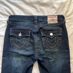 Mörkblå bootcut jeans från True Religion - Säljer ett par mörkblå jeans från True Religion med bootcut passform. Jeansen har dekorativa bakfickor med gula sömmar och silverfärgade knappar. Klassisk femficksmodell och midrise midja! Färgen på jeansen i verkligen är en blandning mellan bild 1 & 2. Jeansen är slitna på båda byxbenen. Annars i jätte bra skick. Nypris: 1299kr. Storlek W29 - Motsvarar 38/M. Byxorna är långa i längden. (Jag är 171 cm och de är för långa för mig).