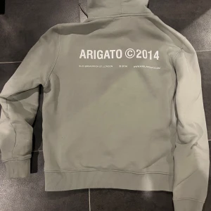 Grå hoodie från Axel Arigato - Säljer en stilren grå hoodie från Axel Arigato med tryck 'ARIGATO ©2014' på både bröstet och ryggen. Tröjan har huva med dragsko och en stor magficka. Perfekt för en avslappnad look. Hoodien är i nyskick och produceras inte längre. 