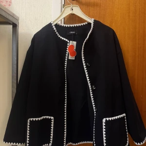 Svart kappa med vita detaljer från Ciler - Svart kappa med rund hals och vita virkade kanter längs öppningen, fickorna och nederkanten. Kappan har stora fickor framtill och knäpps med knappar. Medföljer en matchande halsduk i samma stil.