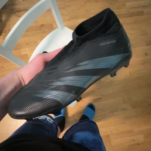 Adidas Predator fotbollsskor i svart och grå - Säljer ett par Adidas Predator fotbollsskor i svart med grå detaljer. Skorna har en strumpliknande passform utan snörning och är tillverkade i syntetmaterial. Perfekta för spel på gräs med sina fasta dobbar och stilrena design.