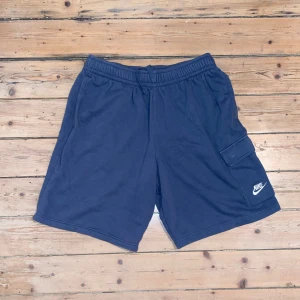 Blå shorts från Nike med cargoficka - Säljer ett par blå Nike shorts med elastisk midja och snörning. De har en praktisk cargoficka på sidan med Nike-logga. Tillverkade i mjuk bomull, perfekta för avslappnade dagar eller träning.