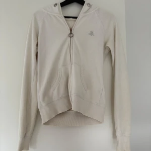 Vit hoodie med dragkedja - Säljer en klassisk vit hoodie med dragkedja och huva. Tröjan har långa ärmar, fickor framtill och dekoreras av ett glittrigt -emblem på bröstet. Ribbstickade muddar och snygg metallring på dragkedjan. Väldigt sliten kofta se bilder men dragkedjan funkar att knäppa o bra skick överlag
