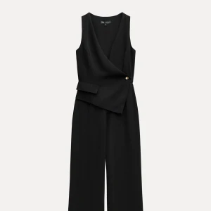 Svart ärmlös jumpsuit med omlott - Snygg svart ärmlös jumpsuit med omlott och v-ringning. Jumpsuiten har en dekorativ knapp i midjan och vida ben. Perfekt för dig som vill ha en stilren och modern look.