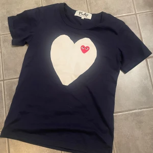 Comme des Garçons T-shirt  - Mörkblå t-shirt från Comme des Garçons Play med stort vitt hjärta och röd hjärt-detalj på bröstet. Klassisk rund halsringning och korta ärmar. Perfekt statement-plagg för en cool och trendig look.