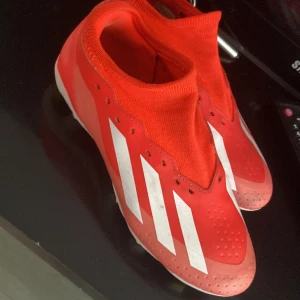 Röda Adidas fotbollsskor Crazyfast - Säljer ett par röda Adidas Crazyfast fotbollsskor med vita ränder och detaljer. Skorna har en strumpliknande passform och plös, samt dobbar i gult och vitt under sulan. Perfekta för dig som vill sticka ut på planen!
