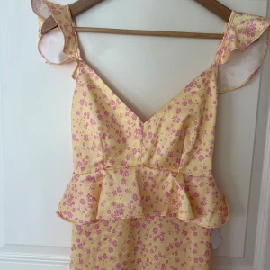 Gul långklänning med volang och blommigt mönster från Andrea Norrman x Bubbleroom  - Söt gul långklänning med rosa blommigt mönster och volangdetaljer. Djup v-ringning, ärmlös med breda axelband och öppen rygg med knyt. Perfekt för sommarens tillfällen.