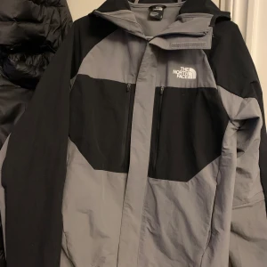 Grå och svart vindjacka från The North Face  - Säljer en grå och svart vindjacka från The North Face använd 1 gång kommer både med dragkedja framtill och två bröstfickor med dragkedja. Jackan har hög krage och logga på bröstet. Perfekt för blåsiga dagar.