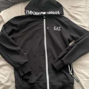 Svart hoodie med dragkedja från Emporio Armani EA7 - Svart hoodie med vit dragkedja och tryckt logga på bröstet samt på huvan. Klassisk passform med långa ärmar och ribbade muddar. Perfekt för en sportig och avslappnad stil. Köpt för 1300