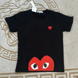 Svart t-shirt med rött hjärta från Comme des Garçons PLAY - Svart t-shirt från Comme des Garçons PLAY med ikoniskt rött hjärta med ögon tryckt både på bröstet och nedtill. Klassisk rund hals och korta ärmar. Perfekt statement-plagg för dig som gillar streetwear.