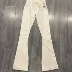 Vita mjukisbyxor från Juicy Couture - Säljer ett par vita mjukisbyxor från Juicy Couture i supermjuk velour. Byxorna har bootcut-modell, dragsko i midjan och ett glittrigt hjärta framtill. På baksidan finns ett broderat motiv med vingar och hjärta i lila och rosa.