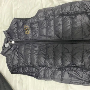 Grå dunväst från EA7 Emporio Armani - Snygg grå dunväst från EA7 Emporio Armani med guldfärgad logga på bröstet. Västen har dragkedja framtill och quiltad design som ger en sportig look. Perfekt att bära över en hoodie eller tröja.