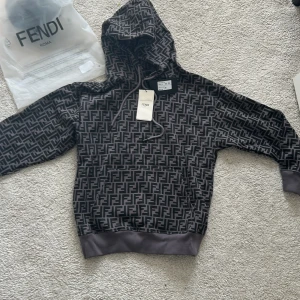 Svart och grå hoodie från Fendi - Säljer en snygg hoodie från Fendi i svart och grått med det klassiska Fendi-mönstret över hela tröjan. Tröjan har huva med dragsko och ribbade muddar vid ärmslut och nederkant. Perfekt för dig som gillar exklusiva streetwear-plagg.