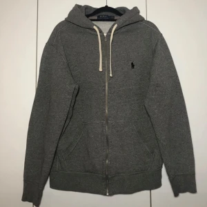 Grå hoodie med dragkedja från Polo Ralph Lauren - Grå hoodie från Polo Ralph Lauren med dragkedja framtill och vit dragsko i huvan. Klassisk logga broderad på bröstet och två fickor framtill. Perfekt för en avslappnad stil.