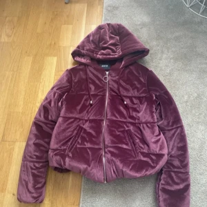 Vinröd pufferjacka med huva från Akoya - Säljer en vinröd pufferjacka från Akoya i mjukt sammetstyg. Jackan har huva, dragkedja framtill och två fickor. Perfekt för kyligare dagar och ger en trendig look.