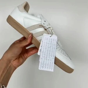 Adidas Samba OG vita sneakers - Säljer ett par Adidas Samba OG sneakers i vitt skinn med beige detaljer och gummisula. Klassisk design med tre ränder på sidan och perforerad tå. Skon har korkinnersula och neutrala färger som passar till mycket.
