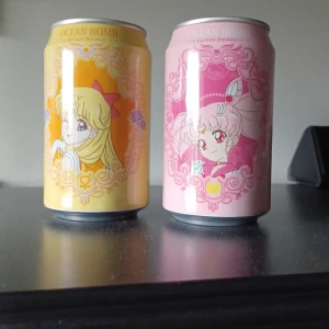 Sailor Moon Ocean Bomb läskburkar - Två samlarburkar med Sailor Moon-motiv från Ocean Bomb. En burk är ljusgul med motiv av Sailor Venus och en är ljusrosa med motiv av Chibiusa. Båda burkarna har dekorativa mönster och japansk text på baksidan. Perfekt för animefans eller samlare.