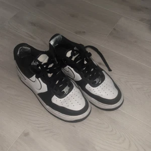 Nike Air Force 1 svartvita sneakers - Säljer ett par klassiska Nike Air Force 1 sneakers i svart och vitt. Skorna har svart ovandel med vita paneler och svart Nike-logga på sidan. De har snörning och perforerad tå för ventilation. Perfekta för dig som gillar streetstyle. Helt okej skick, skador vid båda insida hällarna(bild 5) bild därav pris. Men skadan går säkert att fixa på nått sätt. Finns även ett hål till men ingen större skada (Bild 4). Har ej original packetrt men kan skickas i Nike låda.