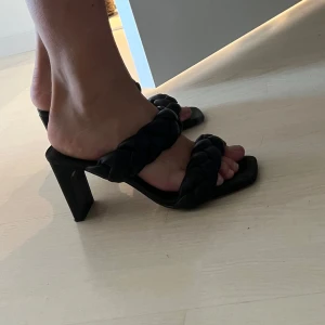 Svarta klackar med flätade remmar - Säljer ett par svarta klackar med fyrkantig tå och breda, flätade remmar. Skorna har en medelhög, kraftig klack och öppen tå, vilket ger en trendig look. Perfekta för dig som vill ha något stilrent men ändå unikt.