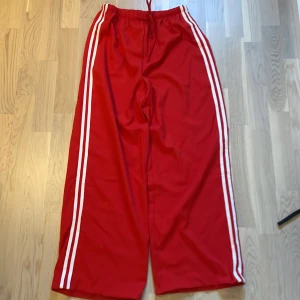 Röda vida träningsbyxor med vita ränder från SHEIN - Röda träningsbyxor från SHEIN med två vita ränder längs sidorna. Vida ben och resår i midjan med dragsko. Tillverkade i 100% polyester, perfekt för en sportig look.