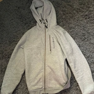 Grå zip från Sail Racing - Säljer en grå hoodie från Sail Racing med dragkedja framtill och en extra dragkedjeficka på bröstet. Tröjan har huva och långa ärmar, perfekt för en avslappnad stil. Loggan är tryckt på bröstet. Hör av dig vid intresse!