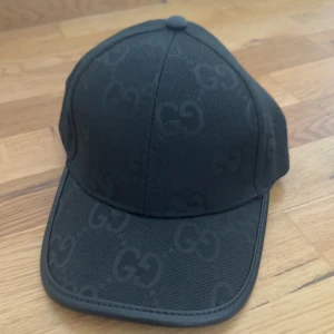 Mörkgrå keps med GG-mönster - Snygg mörkgrå keps med diskret GG-mönster över hela ytan. Klassisk böjd skärm och rundad toppknapp. Perfekt accessoar för en stilren look.