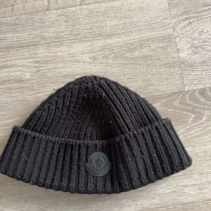 Moncler mössa - Säljer en svart ribbstickad mössa från Moncler med klassisk Moncler-logga framtill. Mössan är i ett tjockare stickat material och har en uppvikt kant för extra stil och värme. Ny pris 3200kr