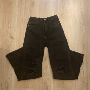 Jeans med slitningar - Svarta jeans med breda ben och slitna detaljer på knäna. Klassisk femficksmodell med knapp och dragkedja i gylfen. De är upp klippta längst ner på benen(men de ska va så). Den ändå defekten är att vissa trådar vid knäna är sönder. Skriv om du har frågor o läs helst min bio innan du köper🫶🏻✨