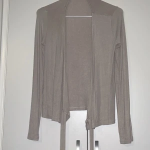 Beige kofta från 157 - Säljer en tunn och mjuk beige kofta från 157 i storlek XS. Koftan har långa ärmar och ett enkelt, öppet snitt med knytband framtill. Perfekt att slänga över en topp för en chill look.