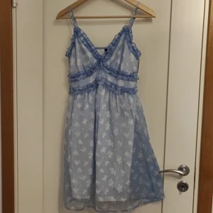 Ljusblå blommig klänning från Vero Moda - Supersöt ljusblå klänning från Vero Moda med vita blommor och volangdetaljer. Klänningen har smala axelband, v-ringning och omlottkänsla framtill. Perfekt för sommarens alla tillfällen!