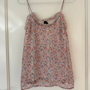 Blommigt linne från Vero Moda - Söt och somrigt linne från Vero Moda i storlek XS. Toppen har smala axelband och ett blommigt mönster i rosa, orange och lila toner på ljus botten. Perfekt för varma dagar och enkel att matcha med jeans eller kjol.