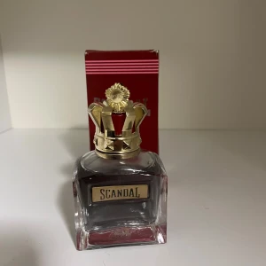 Scandal parfym från Jean Paul Gaultier - En lyxig parfymflaska från Jean Paul Gaultier med namnet Scandal. Flaskan är fyrkantig i genomskinligt glas med en dekorativ guldfärgad krona som lock. Förpackningen är röd med vita ränder och text. Perfekt för dig som gillar unika och eleganta dofter. Knappt använd 