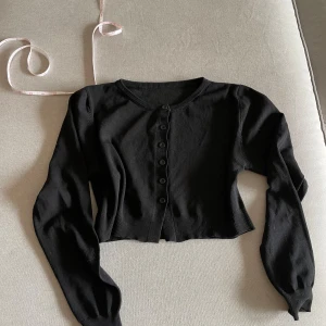 Svart croppad kofta med knappar - Säljer en svart croppad kofta med rund halsringning och långa ärmar. Koftan har ribbade muddar och knäppning framtill med svarta knappar. Perfekt att styla över en topp eller klänning.