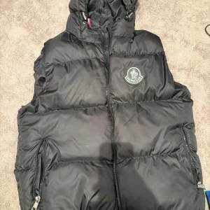 Svart dunväst från Moncler - Säljer en svart dunväst från Moncler med huva och dragkedja framtill. Västen har två fickor med dragkedja och Moncler-logga på bröstet. Perfekt för lager-på-lager och stilren look.