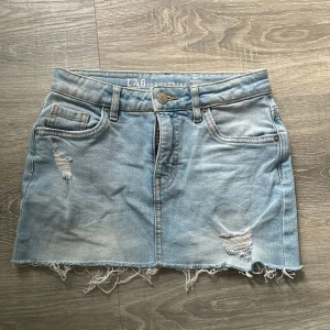 Ljusblå jeanskjol med slitningar från LAB Industries - Trendig ljusblå jeanskjol med slitna detaljer och fransig nederkant. Klassisk femficksmodell med dragkedja och knapp framtill. Perfekt för en avslappnad och cool stil. Säljer för den tyvär är förliten 