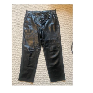 Svarta skinnbyxor från Lindex - Snygga svarta skinnbyxor från Lindex med raka ben och hög midja. Byxorna har dragkedja och knapp framtill samt fickor. Perfekta för dig som vill ha en cool och stilren look.